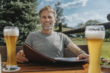 Smiling bearded man holding menu while sitting in beer garden - MFF07645