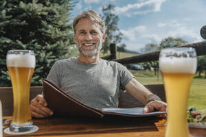 Lächelnder bärtiger Mann mit Speisekarte im Biergarten sitzend - MFF07645