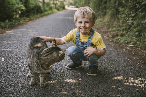 Smiling boy crouching while stroking stray cat on street - MFF07647