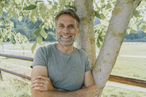Smiling mature man with arms crossed leaning on tree - MFF07653