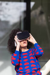 Aufgeregte Frau mit moderner VR-Brille, die virtuelle Realität erlebt, während sie mit erhobenen Armen auf einem Spielplatz Karussell fährt - ADSF22153