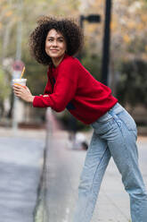 Side view of young African American female standing on street with fresh juice in plastic takeaway cup and enjoying weekend in city - ADSF22154