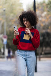 Charmante afroamerikanische Frau mit lockigem Haar und in stilvollem Outfit, die ein Selfie mit einem Plastikbecher mit frischem Saft zum Mitnehmen macht, während sie ihr Smartphone auf der Straße benutzt - ADSF22155
