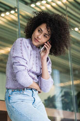 Low angle of African American female with curly hair leaning on glass wall of modern building and speaking on smartphone while looking away - ADSF22159