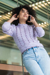 Low angle of African American female with curly hair leaning on glass wall of modern building and speaking on smartphone while looking away - ADSF22160