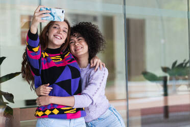 Positive verschiedene Frauen, die sich umarmen und ein Foto mit der Selfie-Kamera eines Smartphones machen, während sie Spaß auf der Straße haben - ADSF22162