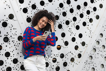 Positive African American female with curly hair and in stylish clothes standing in modern urban district and messaging on smartphone - ADSF22166