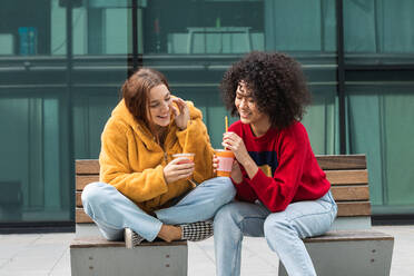 Delighted multiethnic female friends in trendy wear sitting on bench in city and drinking sweet juice to go from plastic cups while looking at each other and spending weekend together - ADSF22167