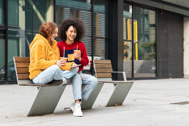 Content multirassischen weiblichen Millennials chatten auf sozialen Medien zusammen auf dem Smartphone, während mit Saft zum Mitnehmen in Plastikbechern auf der Bank auf der Straße sitzen - ADSF22169