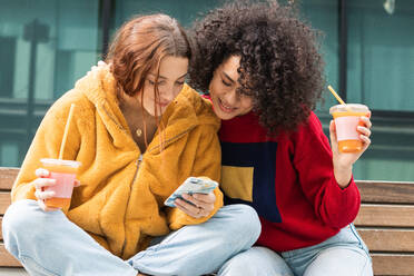 Content multiracial female millennials chatting on social media together on smartphone while sitting with takeaway juice in plastic cups on bench on street - ADSF22170