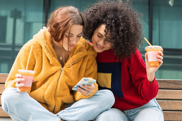 Content multirassischen weiblichen Millennials chatten auf sozialen Medien zusammen auf dem Smartphone, während mit Saft zum Mitnehmen in Plastikbechern auf der Bank auf der Straße sitzen - ADSF22170