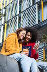 Content multirassischen weiblichen Millennials chatten auf sozialen Medien zusammen auf dem Smartphone, während mit Saft zum Mitnehmen in Plastikbechern auf der Bank auf der Straße sitzen - ADSF22171