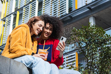 Content multirassischen weiblichen Millennials chatten auf sozialen Medien zusammen auf dem Smartphone, während mit Saft zum Mitnehmen in Plastikbechern auf der Bank auf der Straße sitzen - ADSF22172