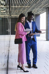 Geschäftsfrau, die ein digitales Tablet benutzt, während sie neben einem Kollegen mit einer Akte im Büroflur steht - PNAF00973