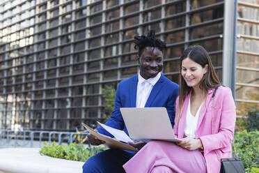 Businesswoman using laptop while sitting by coworker on retaining wall - PNAF00987