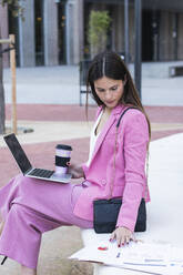 Young female entrepreneur with laptop checking papers while sitting on retaining wall - PNAF00990