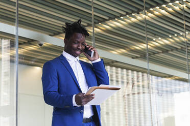 Mittlerer erwachsener Geschäftsmann, der eine Akte überprüft, während er vor einer Glaswand mit einem Mobiltelefon spricht - PNAF01019