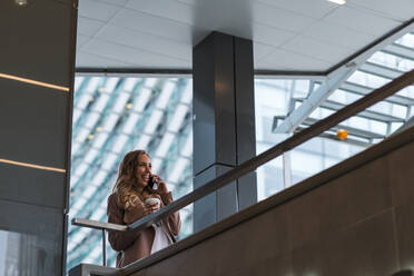 Happy female professional with disposable coffee cup talking on smart phone by railing - JMPF00886