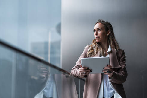 Unternehmerin träumt, während sie ein digitales Tablet vor einer grauen Wand hält - JMPF00895