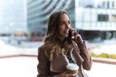 Smiling blond haired businesswoman holding disposable coffee cup while talking on mobile phone - JMPF00898