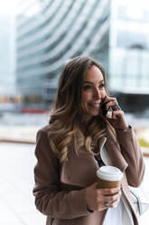 Excited female entrepreneur with coffee cup talking on smart phone while looking away - JMPF00899