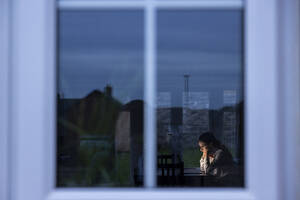 Geschäftsfrau mit Hand am Kinn, die auf einen Laptop schaut, gesehen durch ein Fenster - WPEF04228