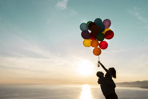 Father carrying daughter playing with bunch of balloons during sunset - LJF02162