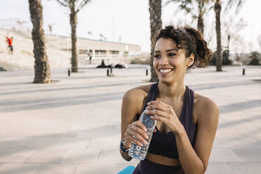 Lächelnde Sportlerin, die mit einer Wasserflasche in der Hand auf dem Fußweg wegschaut - XLGF01357