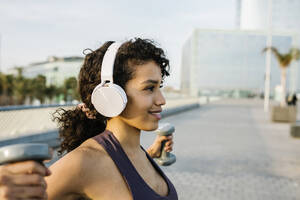 Lächelnde Sportlerin hört Musik über Kopfhörer, während sie mit einer Hantel auf einem Fußweg trainiert - XLGF01368