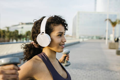 Smiling sportswoman listening music through headphones while exercising with dumbbell on footpath - XLGF01368