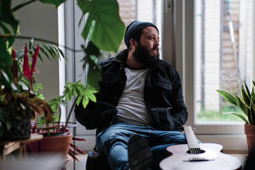 Nachdenklicher Mann sitzt mit Händen in den Taschen am Fenster und Gitarre zu Hause - ASGF00082