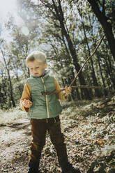 Boy holding stick while standing in forest during sunny day - MFF07663