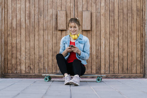 Lächelnde Frau mit Kopfhörern, die ein Smartphone benutzt, während sie auf einem Longboard an einer Holzwand sitzt - EGAF02072