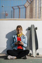 Lächelnde junge Frau mit gekreuzten Beinen, die am Longboard ein Mobiltelefon benutzt - EGAF02074