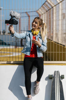 Lächelnde blonde Frau, die ein Video durch eine Kamera aufnimmt, während sie an einer Wand sitzt - EGAF02075