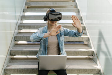 Woman with laptop gesturing while watching video through virtual reality simulator on steps - EGAF02085