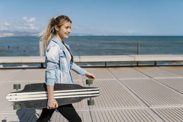 Lächelnde Frau mit Longboard zu Fuß auf dem Pier an einem sonnigen Tag - EGAF02087