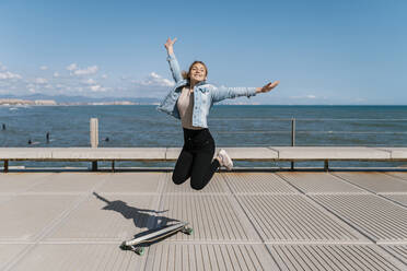 Carefree woman with arms outstretched jumping on pier during sunny day - EGAF02088