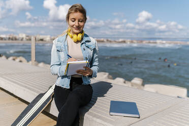 Lächelnde Frau mit Kopfhörern, die in einem Buch schreibt, während sie an einer Mauer sitzt und sich an einem sonnigen Tag zurückhält - EGAF02090