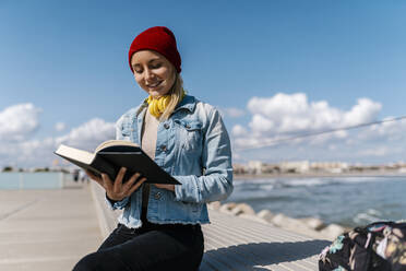 Smiling woman in knit hat reading diary while sitting on retaining wall against sky - EGAF02091