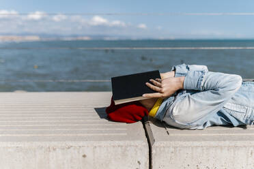 Young woman covering face with diary while lying down on retaining wall during sunny day - EGAF02092