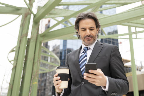 Geschäftsmann, der eine Kaffeetasse hält, während er ein Mobiltelefon gegen eine Struktur benutzt - JCCMF01514