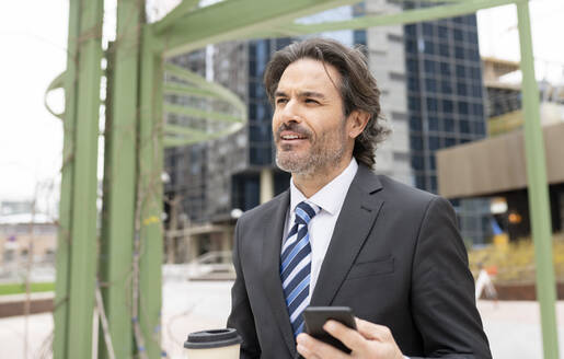 Lächelnder männlicher Fachmann, der wegschaut und eine Kaffeetasse und ein Mobiltelefon hält - JCCMF01517