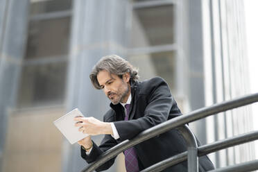 Serious businessman using digital tablet while leaning on railing - JCCMF01538