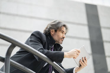 Männlicher Unternehmer träumt, während er ein digitales Tablet am Geländer hält - JCCMF01539