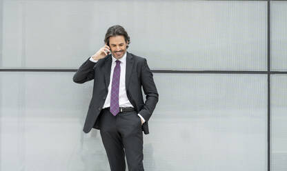 Lächelnder Geschäftsmann mit Hand in der Tasche, der vor einer weißen Wand mit einem Mobiltelefon spricht - JCCMF01540