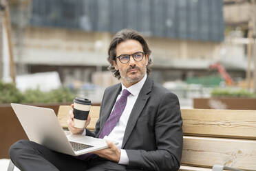 Nachdenklicher Geschäftsmann mit Kaffee in der Hand und Laptop auf einer Bank - JCCMF01546