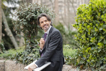 Smiling businessman with laptop looking away while sitting on retaining wall in office park - JCCMF01553