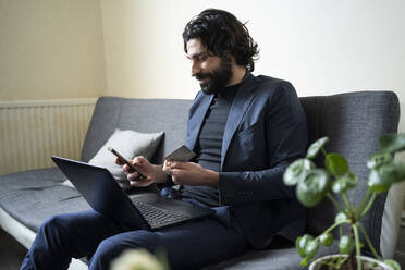 Männlicher Berufstätiger mit Laptop, der ein Smartphone benutzt und eine Kreditkarte zu Hause in der Hand hält - FBAF01626