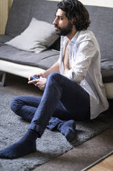 Junger Mann spielt ein Videospiel, während er zu Hause vor dem Sofa sitzt - FBAF01629
